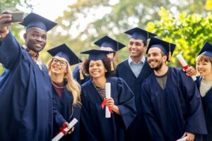 A diverse group of happy international graduates wearing blue gowns and caps taking a selfie outdoors after their graduation ceremony, symbolizing success and global education opportunities in the UK.